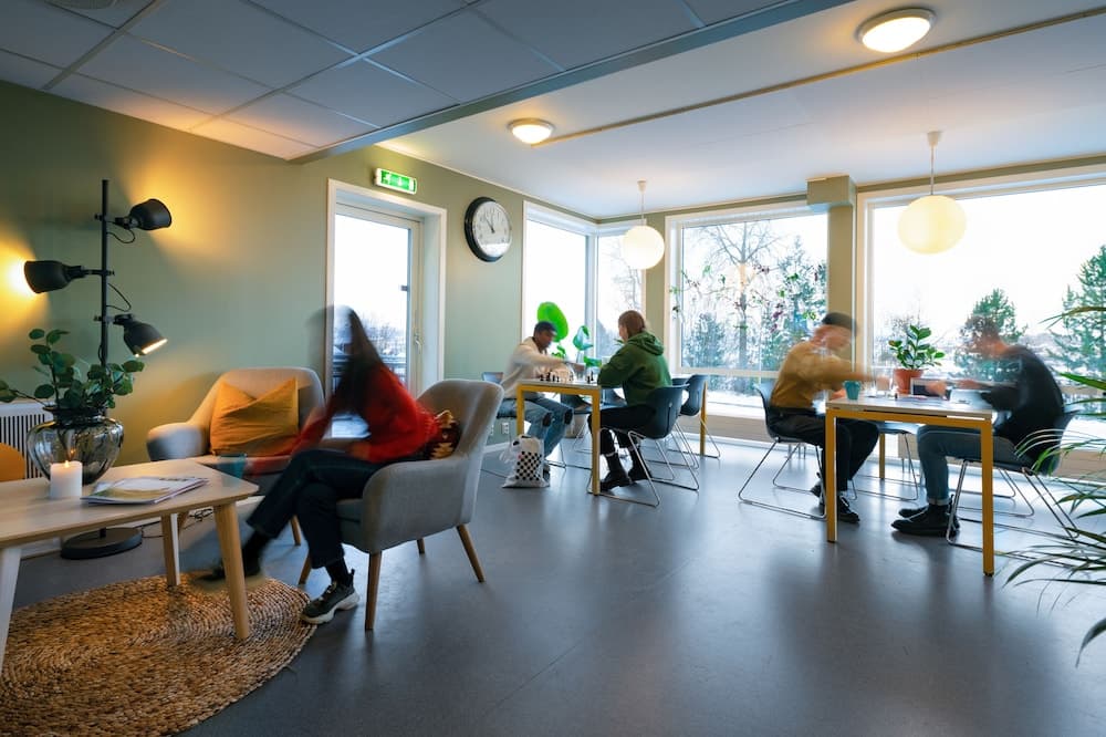 A cozy modern lounge with large windows, warm lighting, and people enjoying games and conversation, ideal for travelers seeking a relaxing break or social interaction in a vibrant indoor setting.