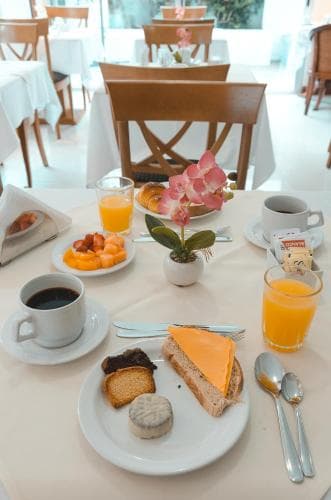 A cozy indoor dining area features a beautifully set breakfast table with coffee, orange juice, pastries, and fresh fruit, creating an inviting atmosphere perfect for travelers seeking a relaxing morning start.
