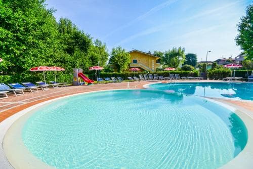 A serene resort pool surrounded by lush greenery, featuring vibrant red umbrellas, lounge chairs, and a playful slide, ideal for family vacations seeking relaxation and fun in a sunny outdoor setting.