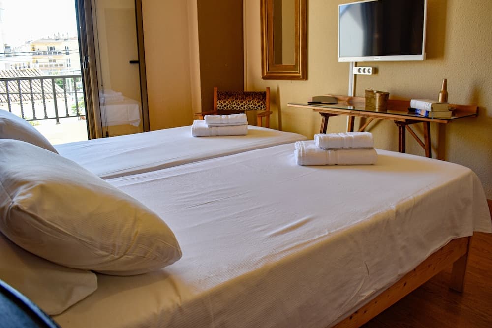 A cozy hotel room with neatly arranged twin beds, a balcony view of buildings, and a wooden desk setup creates a welcoming atmosphere ideal for travelers seeking comfort and convenience.