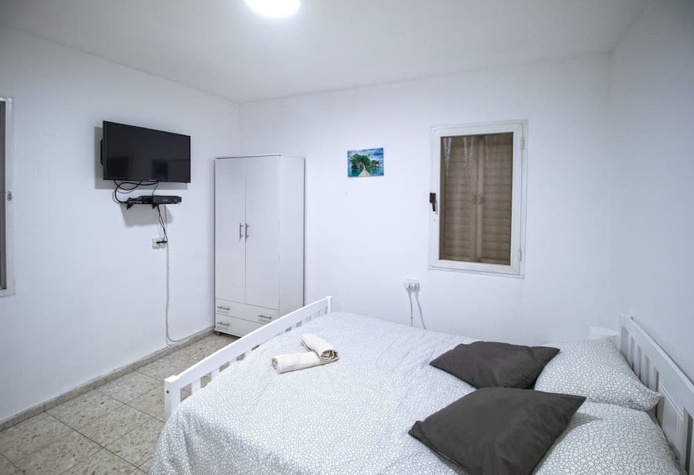 A cozy minimalist bedroom with white walls, a neatly made bed with patterned bedding, a wall-mounted TV, a wardrobe, and a window with blinds, ideal for travelers seeking a clean and simple accommodation option.
