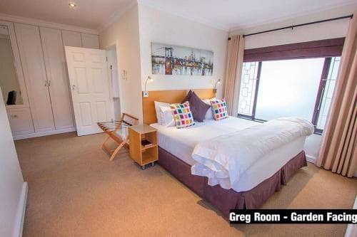 A cozy garden-facing hotel room features a neatly made bed adorned with colorful pillows, a wooden nightstand, and a large window offering a serene view, ideal for travelers seeking comfort and relaxation.