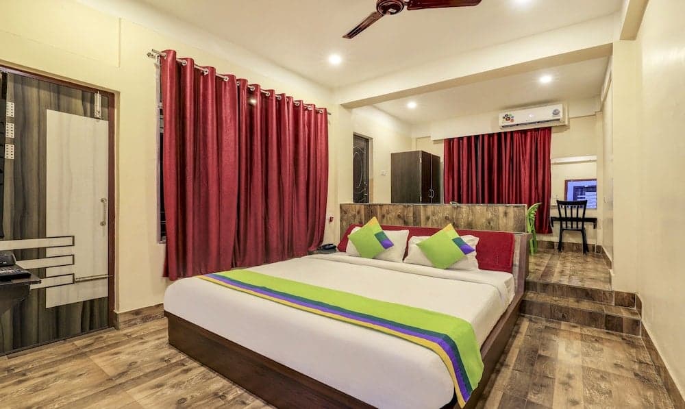 A cozy hotel room features a neatly made bed adorned with colorful pillows and a vibrant throw, set against warm wooden flooring and rich red curtains, creating an inviting atmosphere ideal for travelers seeking comfort and relaxation.