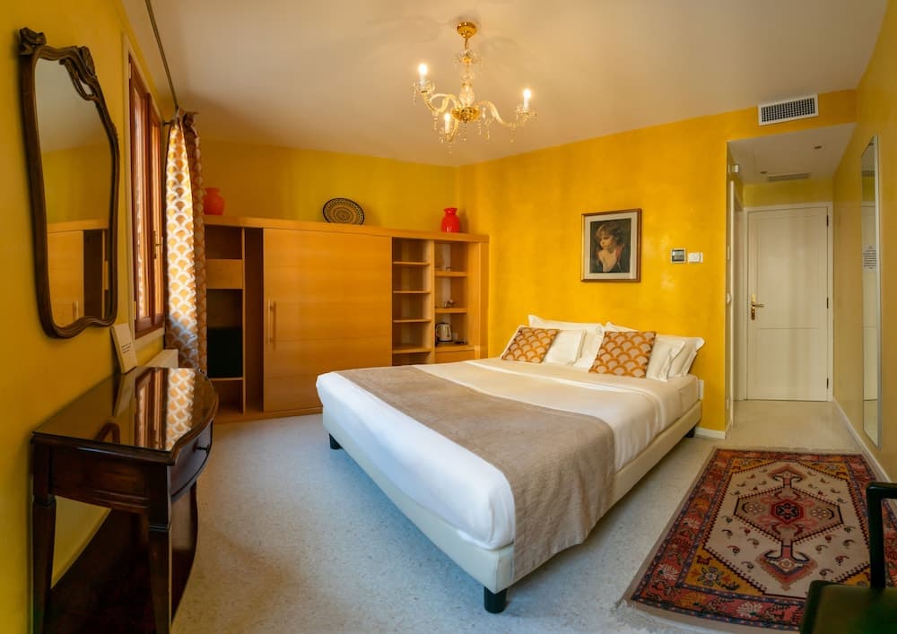 A cozy hotel bedroom with vibrant yellow walls, a neatly made bed, ornate rug, wooden furniture, and a chandelier creates a warm and inviting atmosphere ideal for travelers seeking comfort and style.