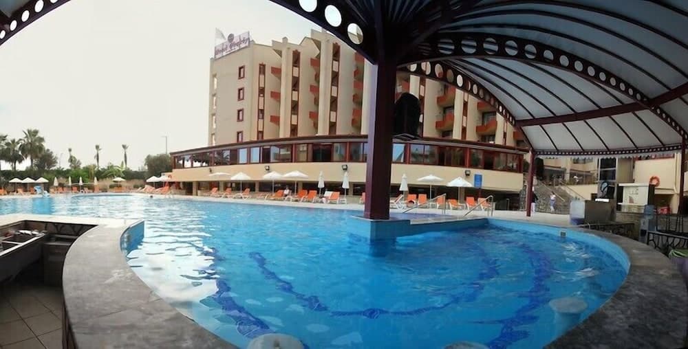 A serene hotel resort scene features a vibrant blue pool under a decorative canopy, surrounded by lounge chairs and umbrellas, with a multi-story building in the background, ideal for vacation relaxation.
