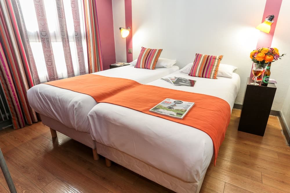 A cozy hotel room with vibrant striped curtains, twin beds adorned with orange throws and striped pillows, and a bedside table featuring flowers and magazines, ideal for travelers seeking comfort and style.