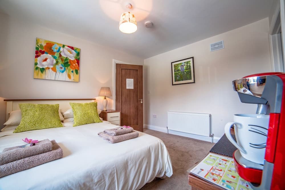 A cozy bedroom with a neatly made bed adorned with green pillows and folded towels, vibrant floral artwork on the walls, a retro red coffee maker on a side table, and soft lighting creates an inviting atmosphere ideal for travelers seeking comfort and relaxation.
