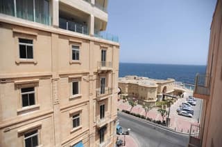 A serene coastal street lined with elegant beige buildings overlooks the calm blue sea, ideal for travelers seeking a luxurious seaside retreat.