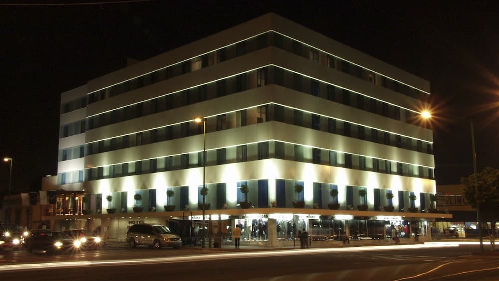 A modern hotel illuminated at night with glowing white lights and potted plants on balconies, creating an inviting urban atmosphere ideal for travelers seeking accommodation in a bustling city setting.