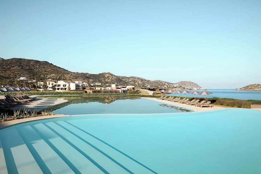 A serene luxury resort features an expansive infinity pool overlooking the calm sea, surrounded by lounge chairs and umbrellas, set against a backdrop of rolling hills under a clear blue sky, ideal for travelers seeking relaxation and scenic beauty.