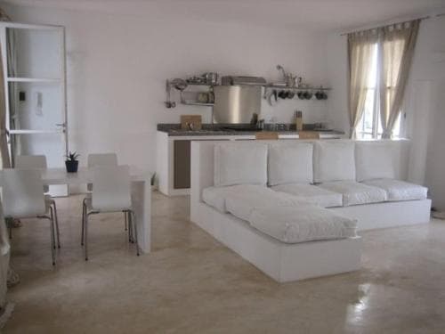 A minimalist apartment interior features a sleek kitchenette, white furniture, and soft natural light filtering through sheer curtains, creating a serene and modern living space ideal for urban retreats.