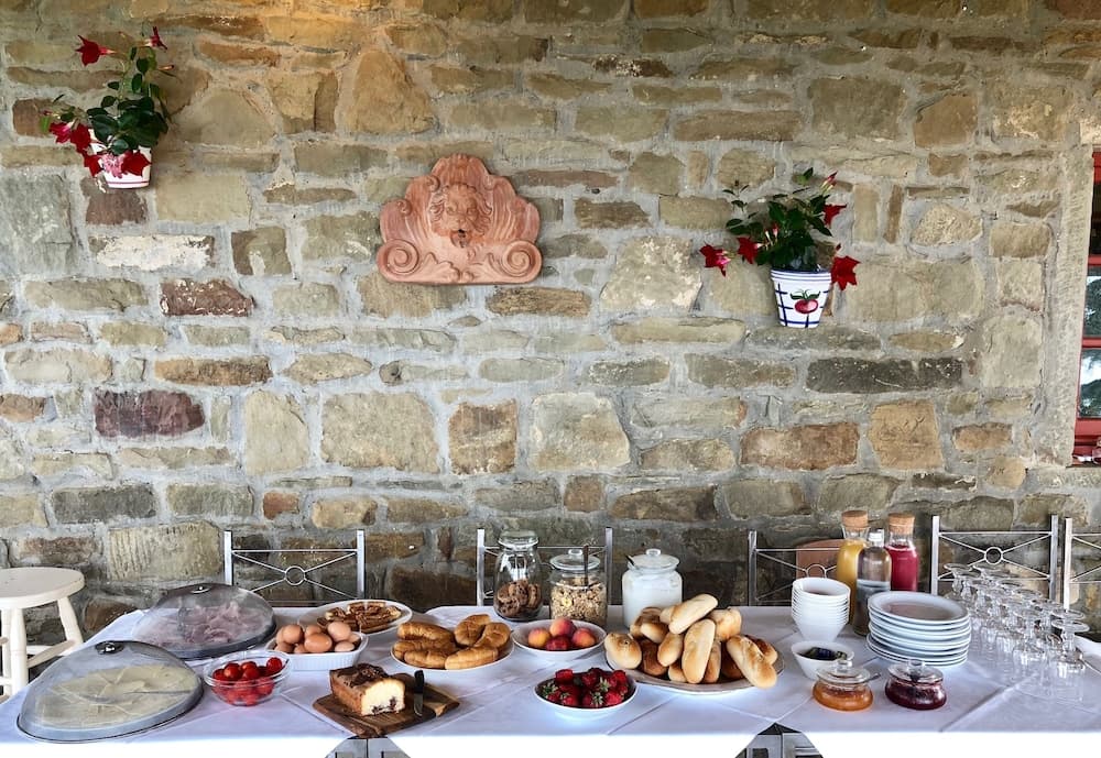 A rustic stone-walled patio adorned with vibrant flowers sets the stage for an inviting breakfast spread featuring fresh breads, fruits, and pastries, perfect for a cozy countryside getaway.