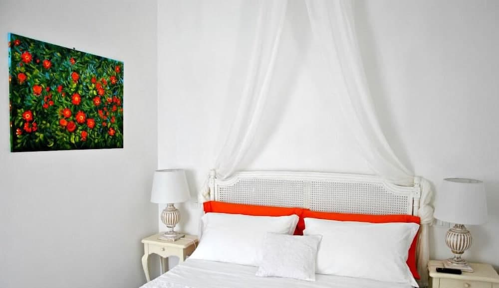 A serene hotel bedroom features a cozy bed with white linens and orange accents under a draped canopy, flanked by elegant bedside lamps and adorned with a vibrant floral painting, ideal for travelers seeking comfort and style.