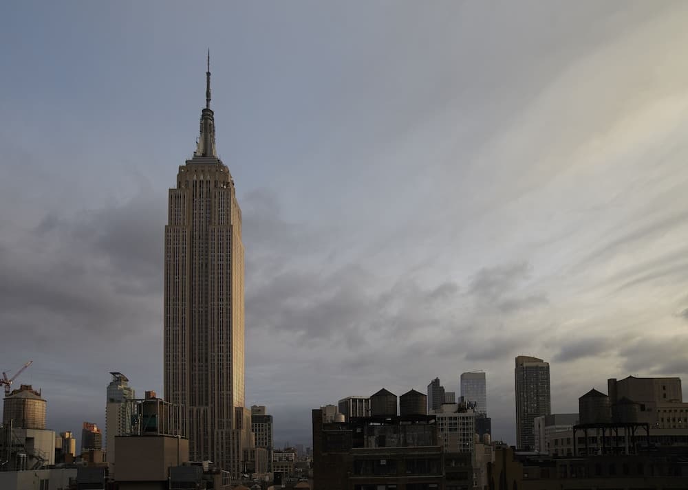 A towering skyscraper dominates the urban skyline under a moody twilight sky, surrounded by industrial rooftops and water tanks, ideal for cityscape photography enthusiasts.
