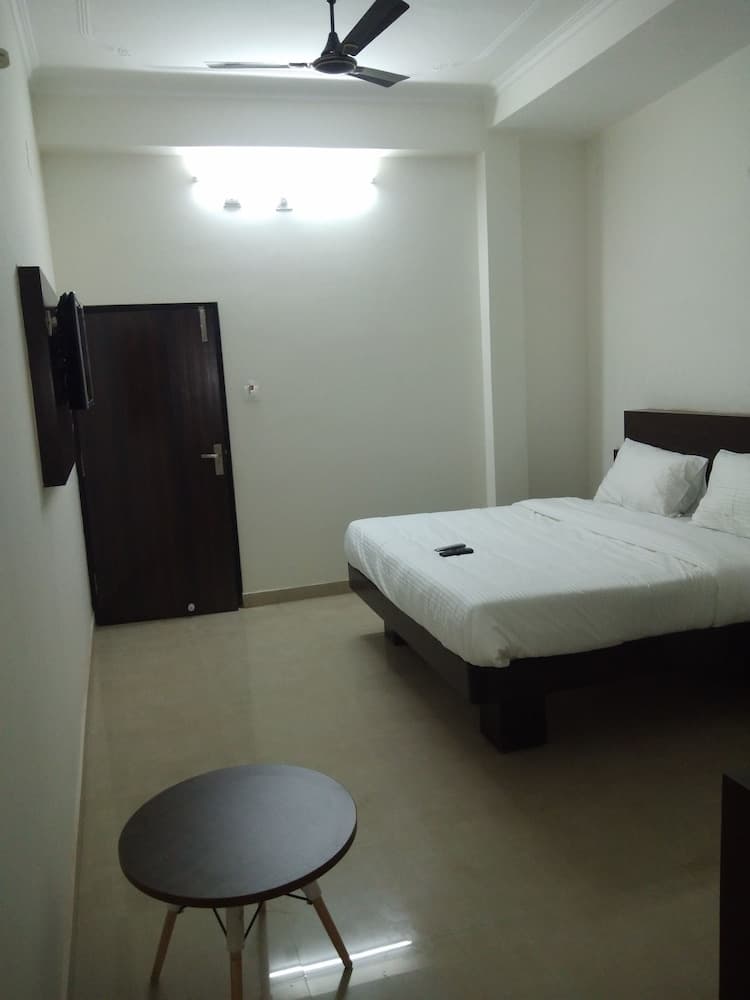 A minimalist hotel room features a neatly made bed, a small round table, a mounted TV, and a ceiling fan, creating a cozy and functional space ideal for travelers seeking simplicity and comfort.