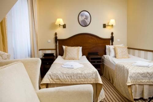 A cozy hotel room features two neatly made beds with wooden headboards, soft lighting from wall sconces, and a comfortable armchair, ideal for travelers seeking a warm and inviting accommodation.