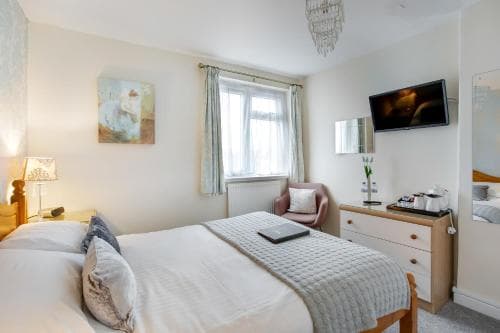 A cozy hotel room features a neatly made bed, soft lighting from a bedside lamp, a comfortable armchair by the window, and modern amenities like a mounted TV, ideal for travelers seeking relaxation and comfort.