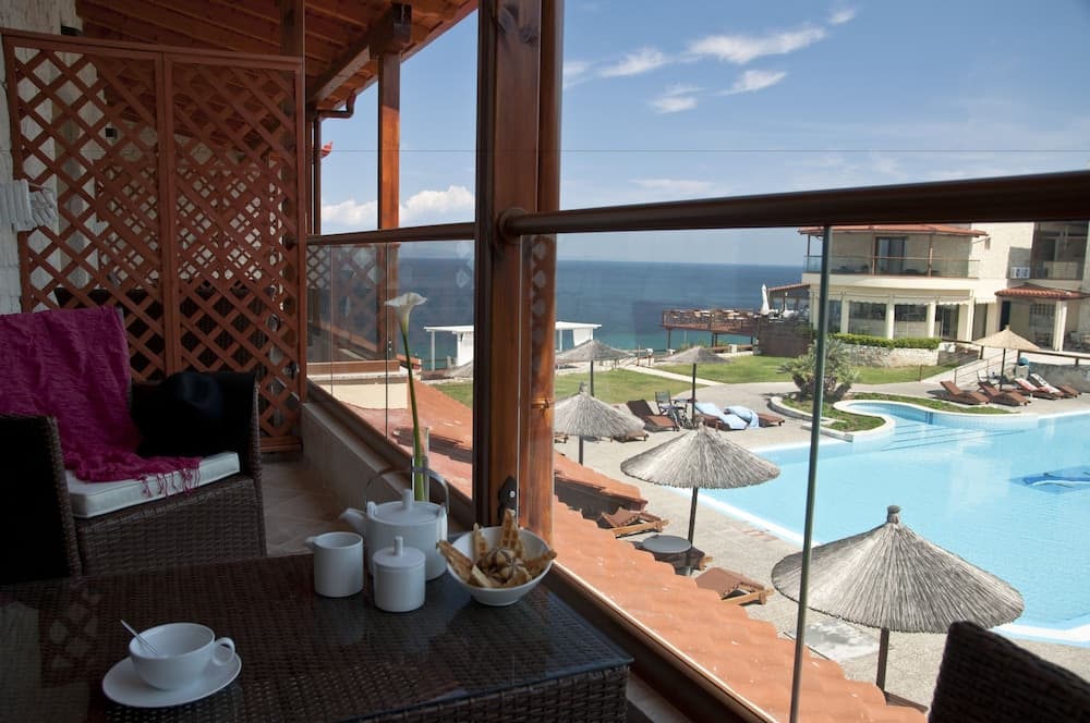 A serene balcony view showcases a cozy breakfast setup overlooking a vibrant poolside area with thatched umbrellas, lush greenery, and the tranquil sea, ideal for a relaxing seaside getaway.