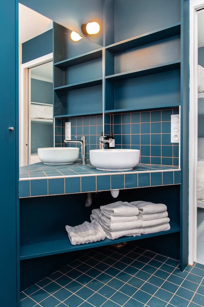 A cozy, modern bathroom features blue tiled walls and floor, dual white sinks, neatly folded towels on shelves, and warm lighting, ideal for travelers seeking stylish and functional accommodations.