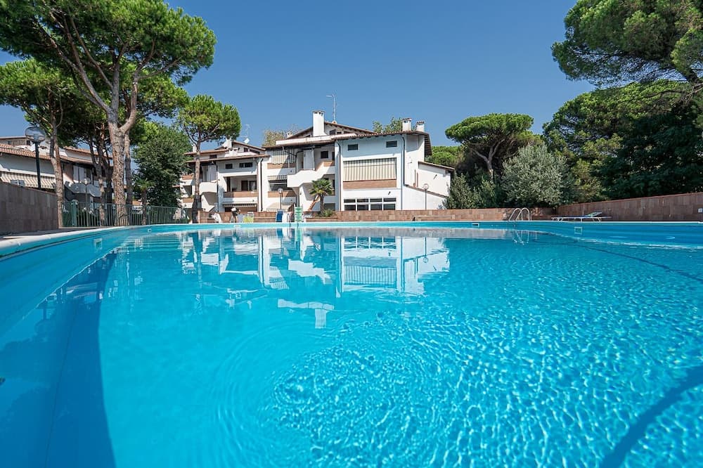 A serene resort pool with crystal-clear blue water reflects the surrounding white buildings and lush green trees under a sunny sky, ideal for vacationers seeking relaxation and leisure.