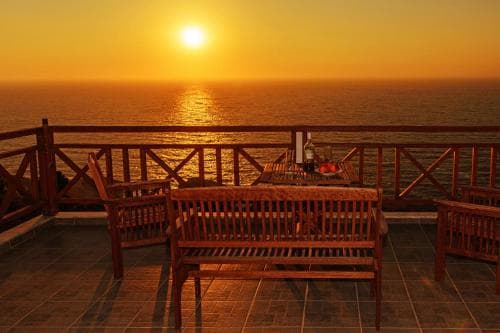 A serene seaside terrace at sunset features wooden benches and tables, perfect for relaxing and enjoying the tranquil ocean view, ideal for travel enthusiasts seeking peaceful coastal retreats.