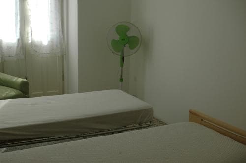 A cozy, minimalist bedroom with two simple beds, a green fan, and soft natural light filtering through sheer curtains, ideal for budget travelers seeking comfort in a serene setting.