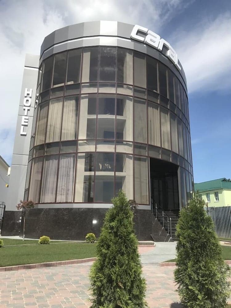 A modern cylindrical hotel with large glass windows and a "Carat" sign stands under a bright blue sky, surrounded by neatly trimmed shrubs, ideal for travelers seeking upscale accommodations in an urban setting.
