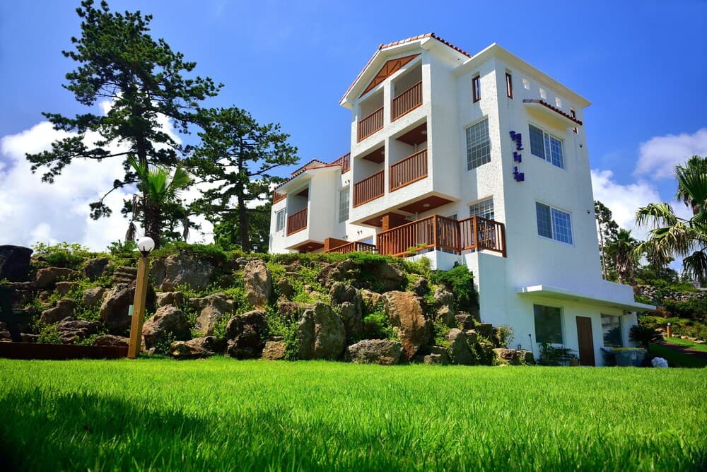 A serene coastal villa with white walls, wooden balconies, and lush greenery offers a picturesque retreat under a clear blue sky, ideal for luxury vacation stays.