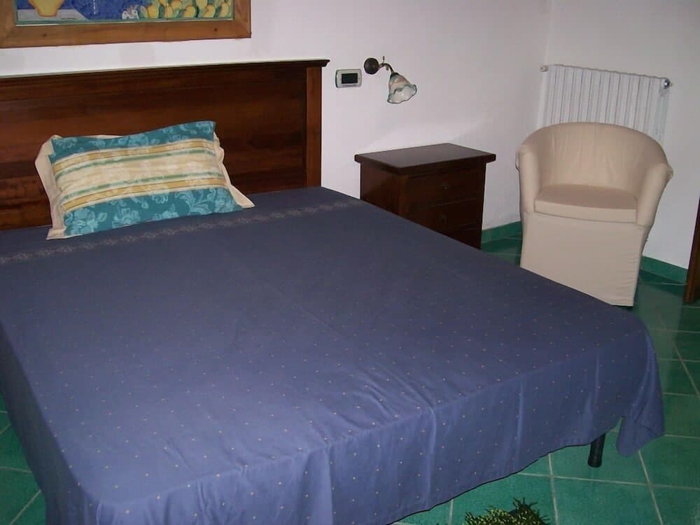 A cozy bedroom with a neatly made blue bed, wooden furniture, a beige armchair, and green tiled flooring offers a serene retreat for travelers seeking comfort and relaxation.