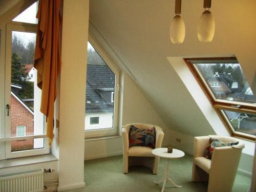 A cozy attic lounge with slanted ceilings, comfortable armchairs, and large windows offering a serene view of neighboring houses, perfect for a tranquil getaway or vacation rental experience.