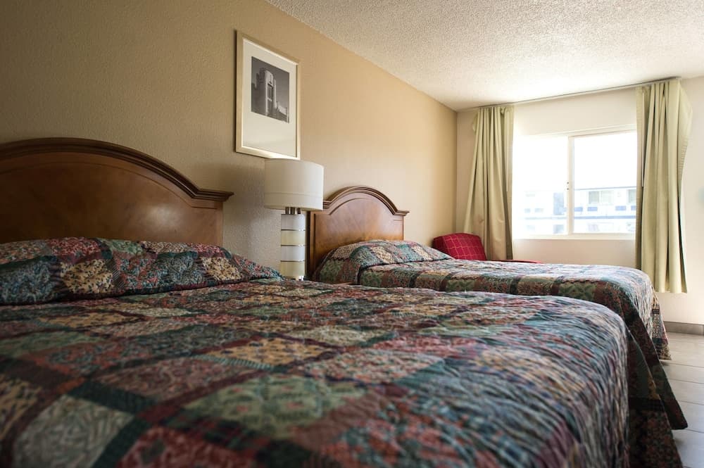 A cozy motel room features two neatly made beds with patterned quilts, wooden headboards, a framed black-and-white photo on the wall, and a window with beige curtains, ideal for travelers seeking comfortable accommodations.
