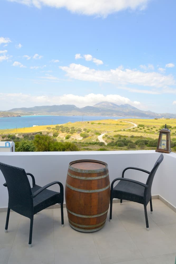 A serene balcony with two chairs and a wooden barrel overlooks a picturesque coastal landscape under a bright blue sky, ideal for a relaxing getaway or vacation rental promotion.