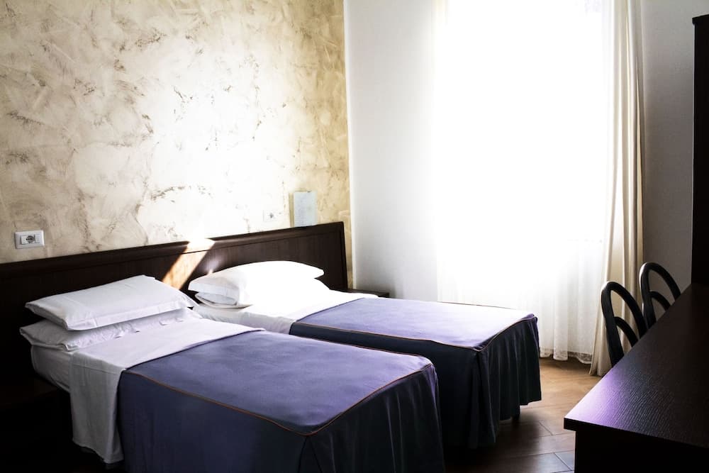 A cozy hotel room features two neatly made beds with purple bedspreads, a desk by the window with sheer curtains, and a textured wall, ideal for travelers seeking comfort and simplicity.