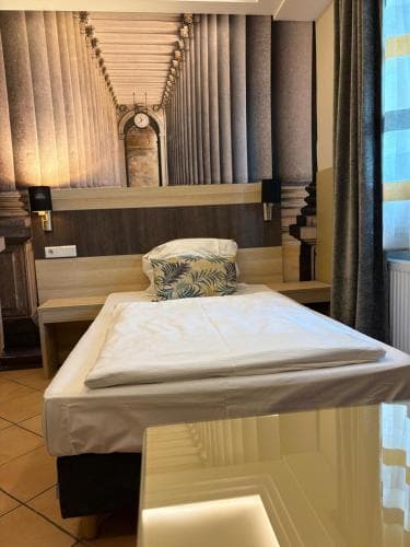 A cozy hotel room features a neatly made bed with a tropical-patterned pillow against a wall mural of classical columns, creating an inviting atmosphere ideal for travelers seeking comfort and cultural ambiance.