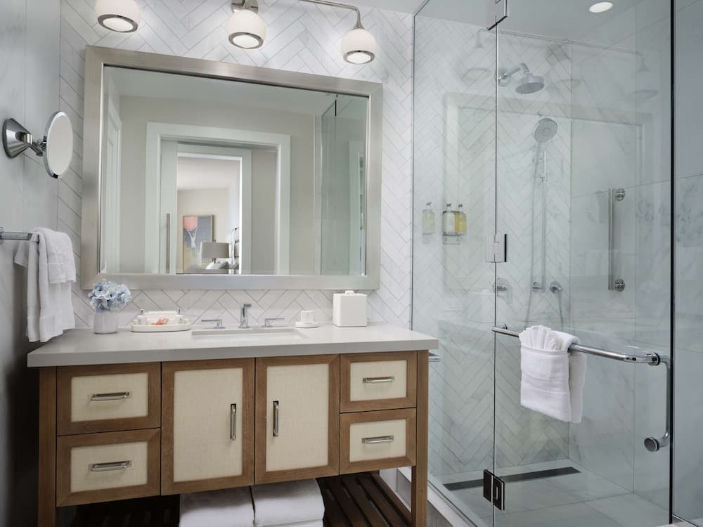 A serene hotel bathroom features a modern vanity with dual sinks, a sleek glass-enclosed shower, and herringbone tile walls, ideal for travelers seeking comfort and style.