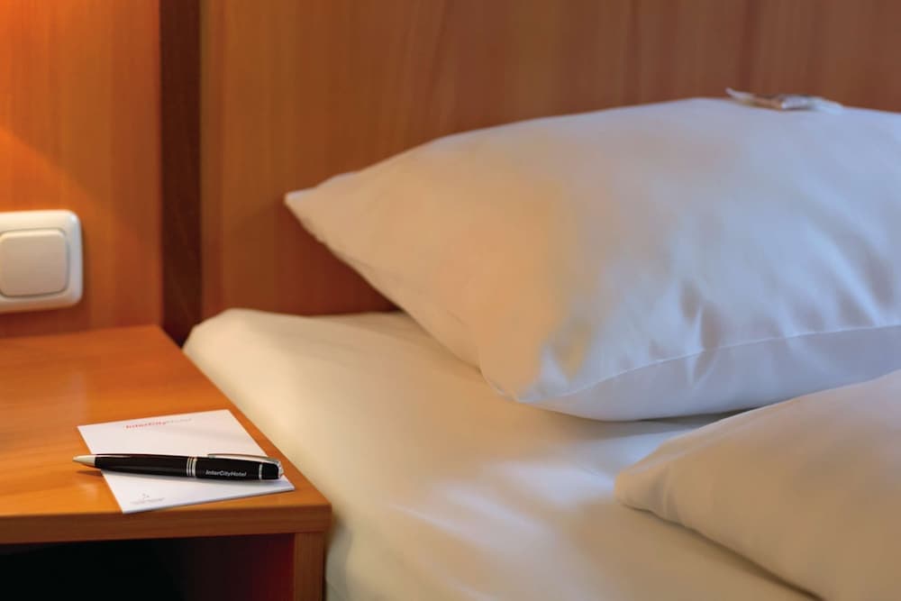 A cozy hotel room corner featuring a neatly made bed with white linens, a wooden bedside table holding a pen and notepad, evokes a serene and inviting atmosphere ideal for travelers seeking comfort and convenience.