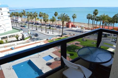 A serene coastal balcony view showcases a swimming pool, palm-lined promenade, and the tranquil sea, ideal for relaxing seaside getaways.