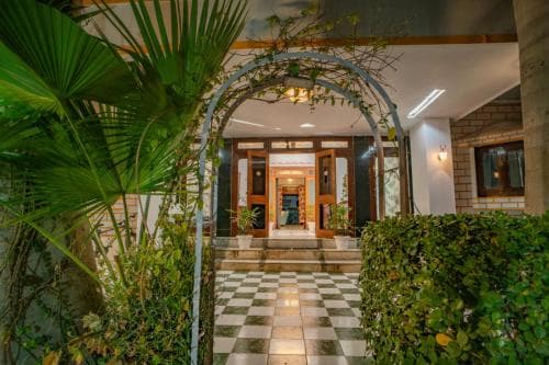 A serene tropical resort entrance features a checkered pathway leading through lush greenery under a vine-covered archway, ideal for travelers seeking a tranquil getaway.