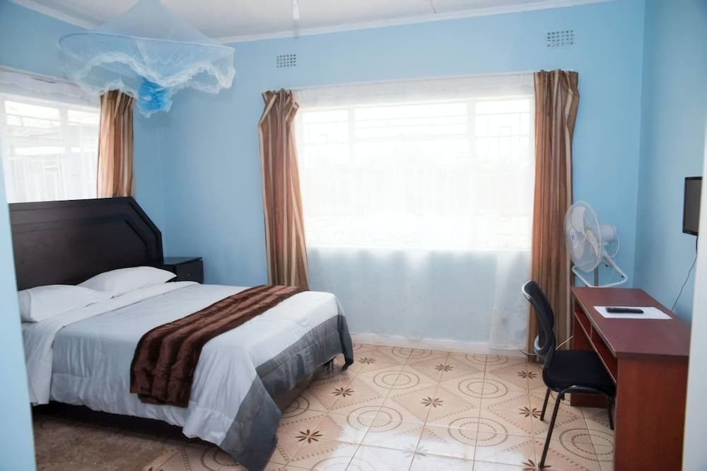 A serene hotel room with light blue walls, a cozy bed adorned with a mosquito net, a tiled floor with floral patterns, and a desk with a fan, ideal for travelers seeking comfort and relaxation.