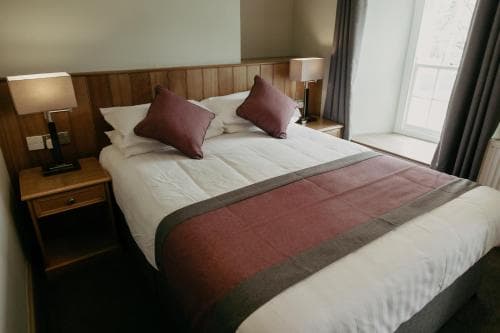 A cozy hotel room features a neatly made bed with white linens and purple accents, wooden bedside tables with lamps, and a large window letting in natural light, ideal for travelers seeking comfort and relaxation.
