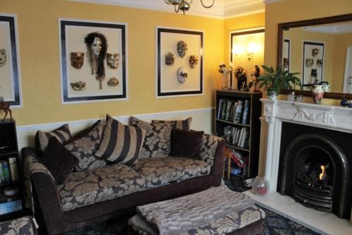 A cozy living room with warm yellow walls, framed artwork, a plush patterned sofa, and a lit fireplace creates an inviting atmosphere perfect for a relaxing getaway or home stay experience.