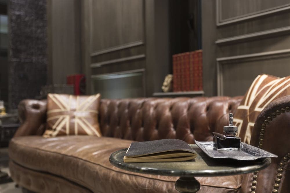 A cozy lounge area features a plush leather sofa adorned with Union Jack pillows, a vintage book, and a cigar on a tray, creating an inviting atmosphere ideal for travelers seeking relaxation in a boutique hotel setting.