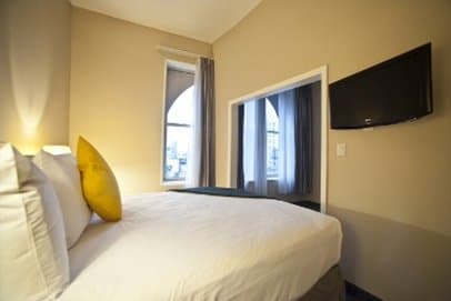 A cozy hotel room with soft lighting, a neatly made bed adorned with a yellow pillow, large windows draped in curtains, and a wall-mounted TV, ideal for travelers seeking comfort and relaxation.