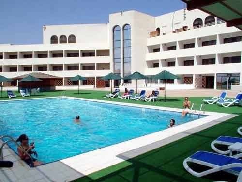 A vibrant resort scene features a large swimming pool surrounded by lounge chairs and umbrellas, set against a multi-story building under a clear blue sky, ideal for travelers seeking relaxation and leisure.