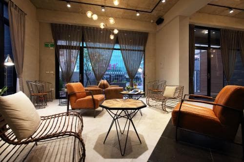 A cozy modern lounge with orange armchairs, wicker seating, and large windows draped in sheer curtains creates an inviting atmosphere ideal for travelers seeking relaxation or a quiet workspace.