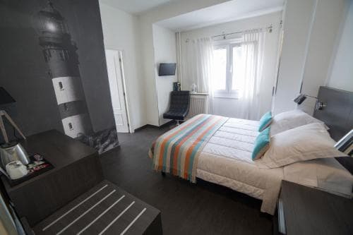 A cozy hotel room features a neatly made bed with colorful bedding, a lighthouse mural on the wall, modern furnishings, and soft natural light from a window, ideal for travelers seeking comfort and style.
