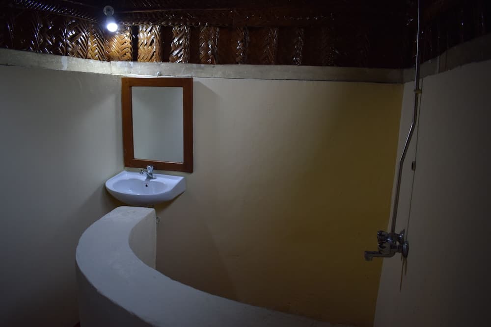 A cozy bathroom with a rustic thatched ceiling, white sink, wooden-framed mirror, and curved bench, ideal for travelers seeking an eco-friendly accommodation experience.