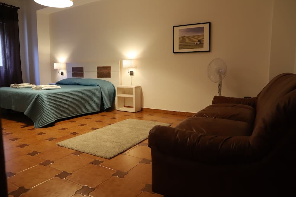 A cozy hotel room features a neatly made bed with teal bedding, a brown leather sofa, terracotta-tiled flooring, and warm lighting, ideal for travelers seeking comfort and relaxation.
