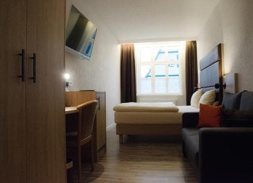 A cozy hotel room features a neatly made bed by a bright window, wooden furnishings, and warm lighting, ideal for travelers seeking comfort and simplicity.