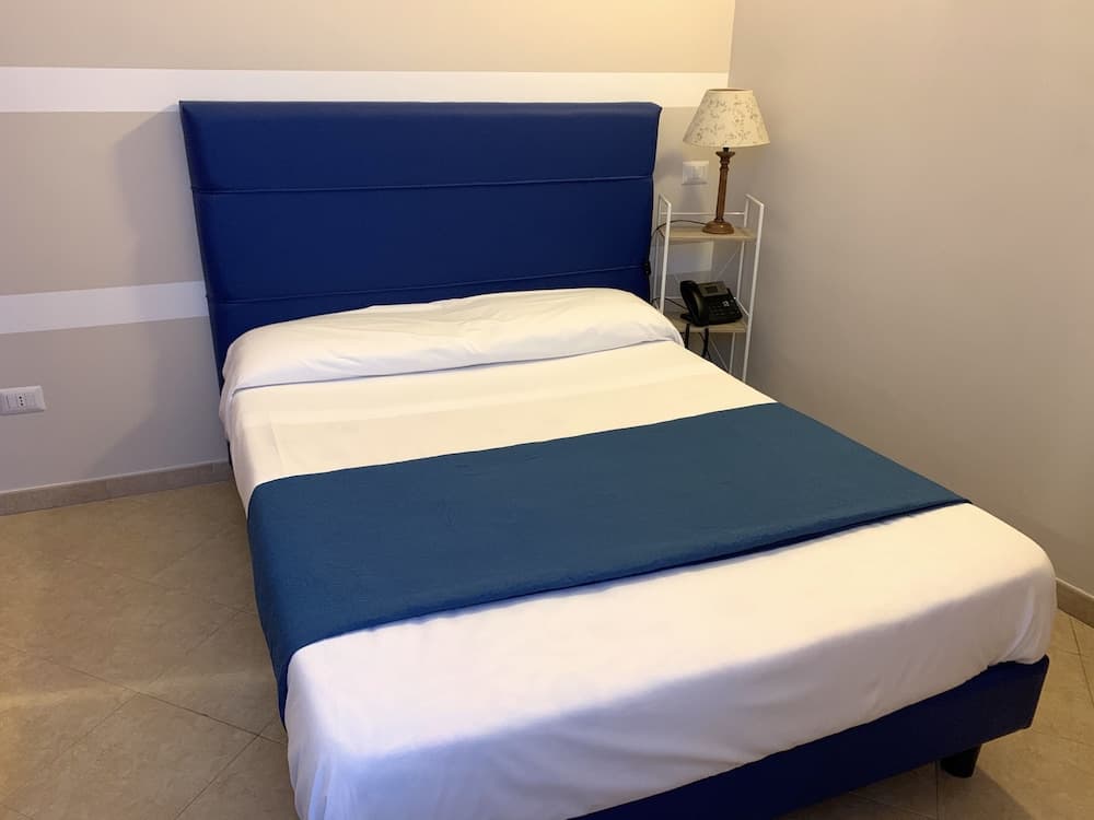 A cozy hotel room features a neatly made bed with blue and white linens, a bedside lamp, and a phone on a small shelf, ideal for travelers seeking comfort and simplicity.